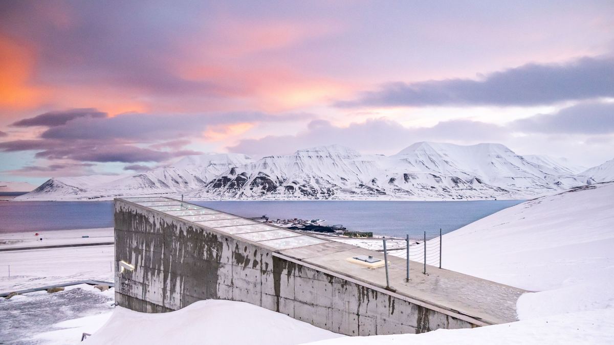 A weathered concrete building with a angled roof stands on a snowy slope above calm water, with snowy mountains and a pink and purple sunset sky behind.